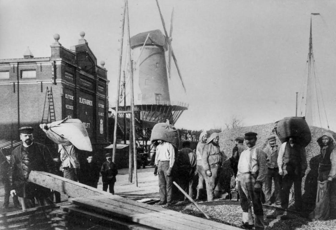 6, Molen 03504 a Het Fortuin / Slikmolen / Molen van Rossum (Delft), foto onbekend, opname circa 1906
&nbsp;Calvé&nbsp;Zakkendragers bezig met het lossen van een schip voor de fabrieken van &nbsp;de Nederlandsche Oliefabriek (NOF) van Calvé aan de Wateringseweg met op de achtergrond een deel van de fabrieksgebouwen en de molen Het Fortuin. Links met pet staat meester C.G. van den Broek. | Database Nederlandse molens