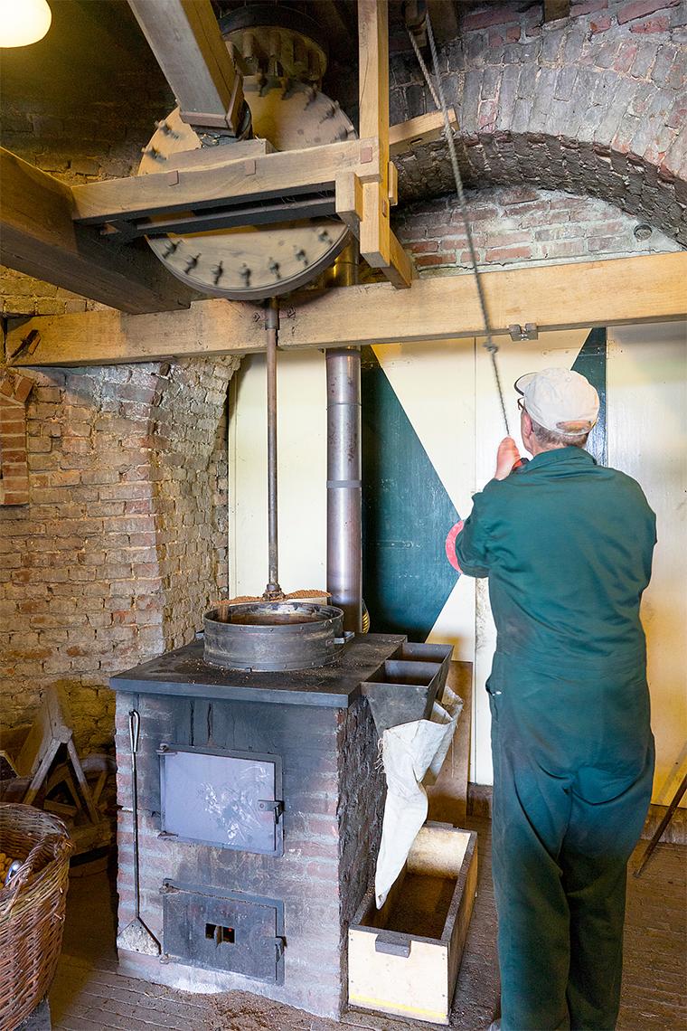 15, Molen 02781 Windlust (Nederweert-Roeven), Marcel van Nies (25-4-2018)Op de houtgestookte vuister wordt het meel verwarmd. Het roerijzer zorgt ervoor dat het gelijkmatig gebeurt, en voorkomt aanbranden. De olieslager haalt het roerijzer omhoog om de bulen te kunnen vullen. | Database Nederlandse molens