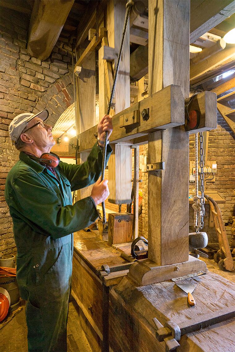 18, Molen 02781 Windlust (Nederweert-Roeven), Marcel van Nies (25-4-2018)Met de slaghei wordt de slagbeitel (een wig) aangeslagen. De druk in de perslade, die bij elke slag toeneemt, perst de olie uit het warme meel. De olie druppelt onder de slagbank in een pan. | Database Nederlandse molens