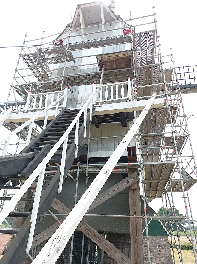 12, Molen 12350 Aurora (Baexem), Frans Verstappen (18-6-2023)Op de Aurora molen In Baexem werden de zijwegen van nieuw kleedhout voorzien. Bij het vervangen van de planken werd geconstateerd dat de kopse kanten van de steenbalk aangetast waren door de knaagkever. Een gespecialiseerd bedrijf moest er aan te pas komen om de steenbalk te conserveren. Had men vroeger slechts een spoorstijl in het midden van de steenbalk, naderhand werden dat er twee. Dit om te voorkomen dat de kast de neiging zou krijgen in te zakken. Deze steenbalk had ooit een spoorstijl die later aangepast werd naar twee, een historische balk dus. De steenbalk is nu gerestaureerd en wat belangrijk is, dat hij behouden is. De Aurora molen is enkele centimeters opgevijzeld en staat dus op een bok. Op deze manier is de druk op de steenbalk overgenomen door de potkrikken die onder de hoekstijlen staan. De molen staat in de steiger omdat het kleedhout vervangen moet worden. | Database Nederlandse molens